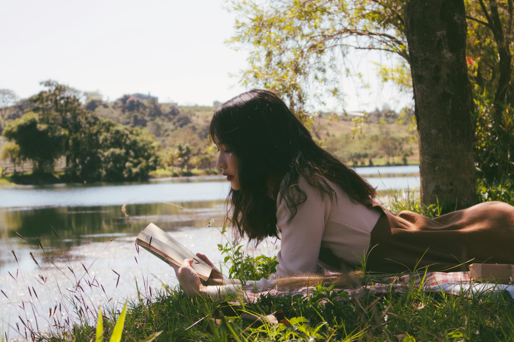 A woman reading a book on the grass beside a sunlit lake.